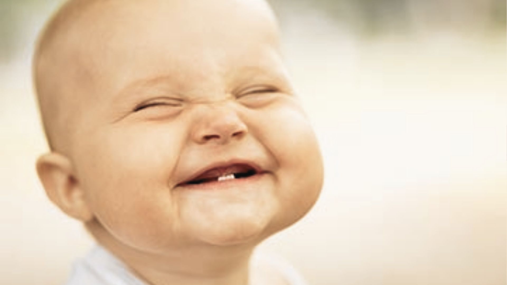 Joyful baby scrunching eyes shut while laughing, revealing two tiny bottom front teeth.