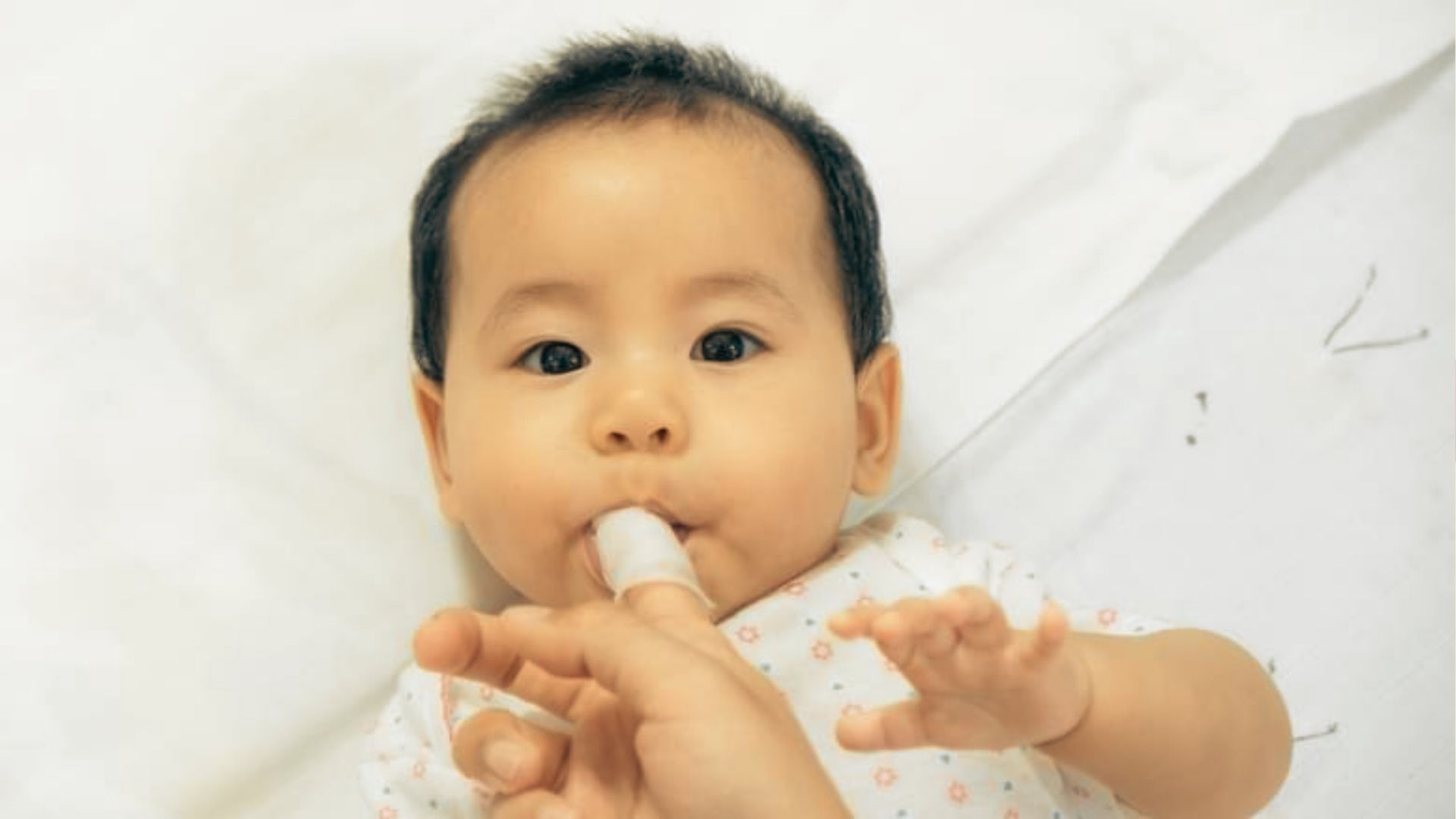 A baby lies on a white sheet while an adult gently cleans inside the baby’s mouth with a gauze-wrapped finger.