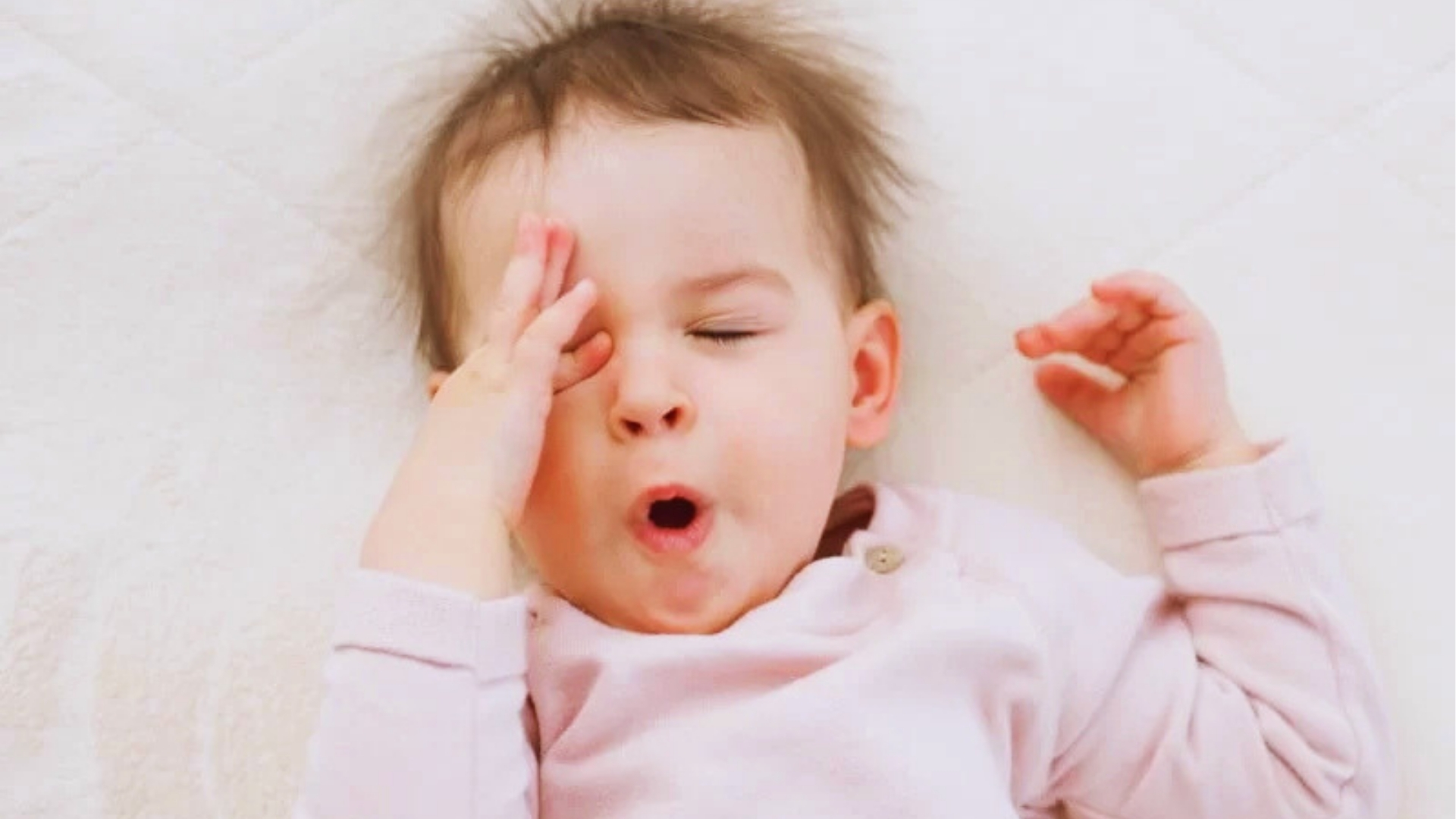 Sleepy baby in pink rubbing eye and yawning, since rapid head shaking often signals tiredness or overstimulation in infants.