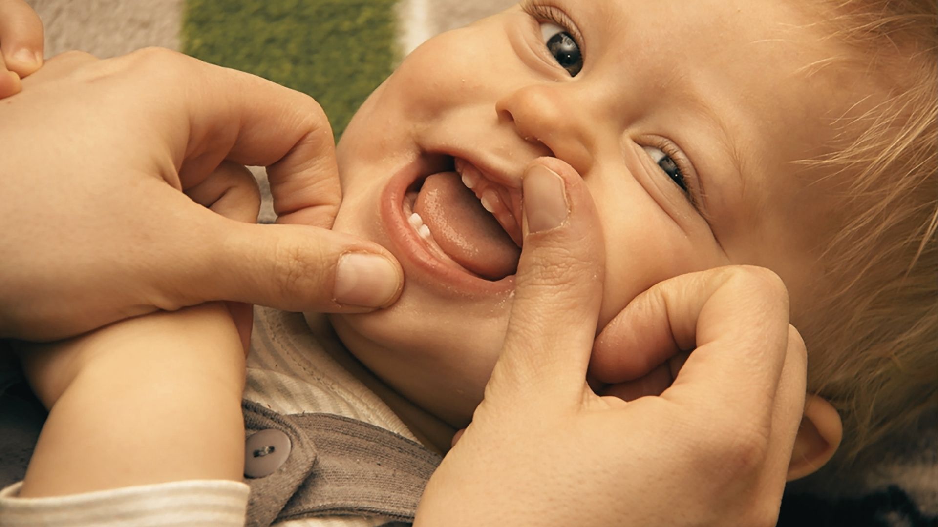 An adult’s hands gently pull back a smiling toddler’s lips to show the inside of the mouth.