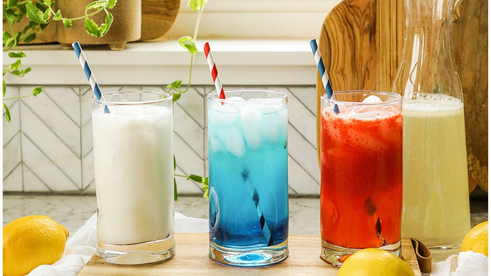 Three tall glasses with red, white, and blue drinks sit on a wooden board with striped paper straws and fresh lemons.