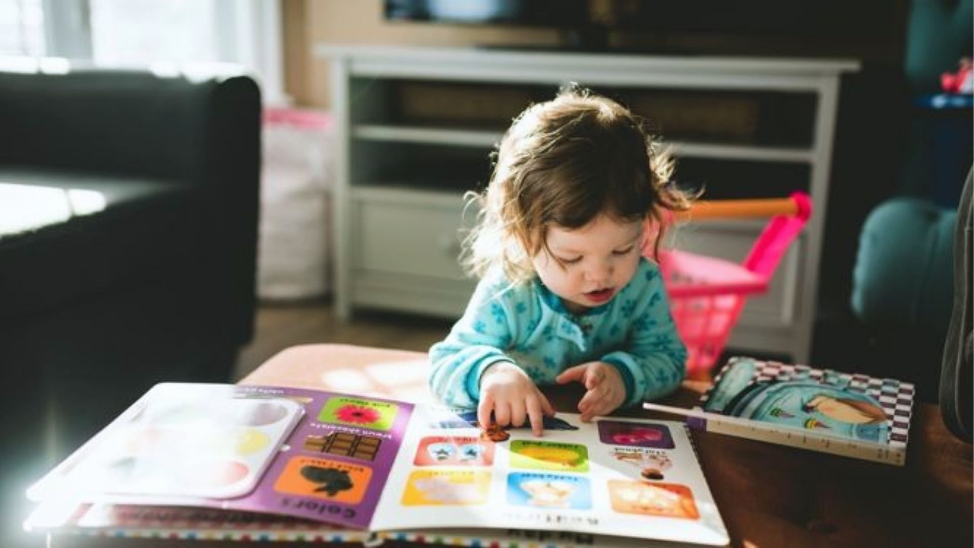 Toddler in teal pajamas leans over a coffee table, pointing to a colorful picture book in a sunlit living room.