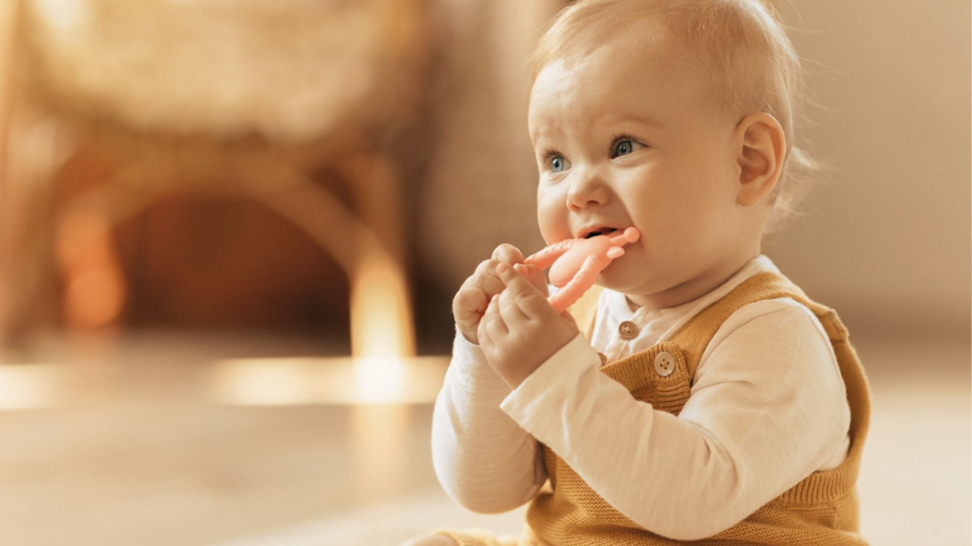 Baby wearing a knit vest sits on the floor, biting a pink hand-shaped teether toy.